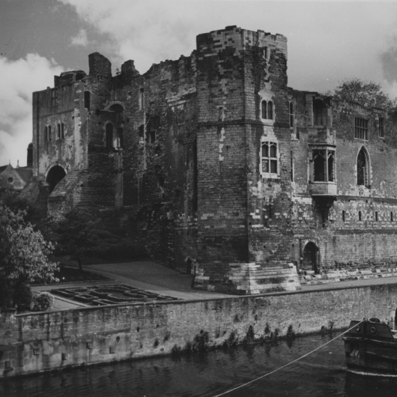 “Historic stone castle with tall walls and arched windows beside a river, with a barge moored in the foreground.