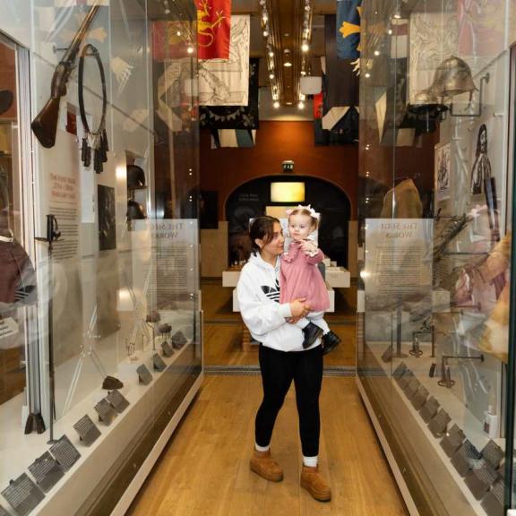Woman holds small female child as they walk between the glass display cases at the museum