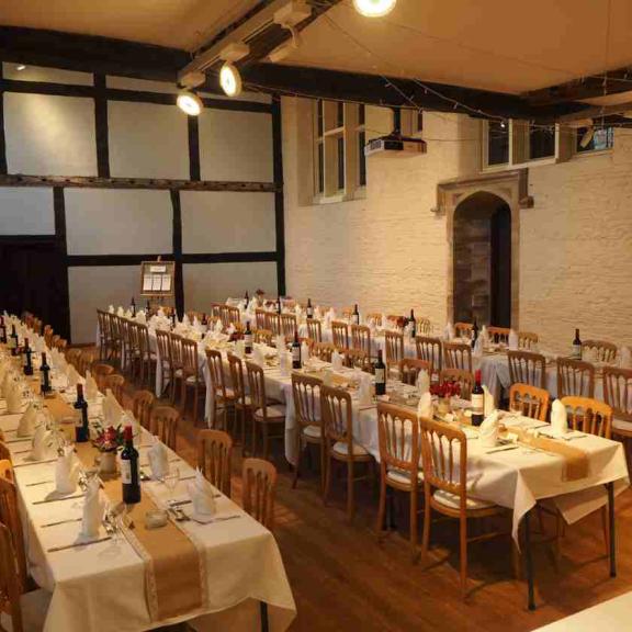 Long banquet tables set for a formal event in a historic hall with wooden beams and white brick walls, featuring neatly arranged chairs, white tablecloths, folded napkins, wine bottles, and floral centrepieces