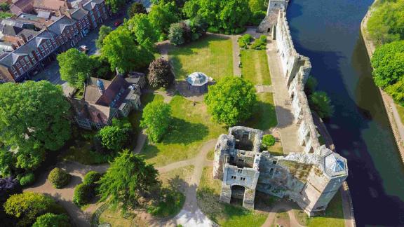 Aerial view of Newark Castle ruins in Nottinghamshire, England, showing stone walls along the River Trent, green lawns, mature trees, and nearby town buildings.
