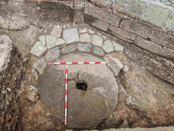 Large worn millstone covering a medieval cesspit, surrounded by stonework, with measuring rods placed across the surface.