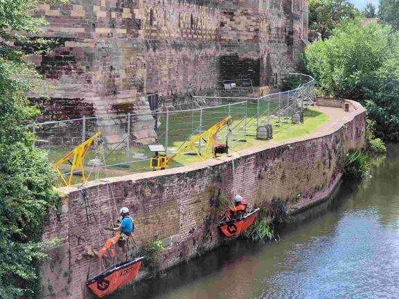 Masons suspended over the river repair Newark Castle’s riverside wall using ropes and safety gear.