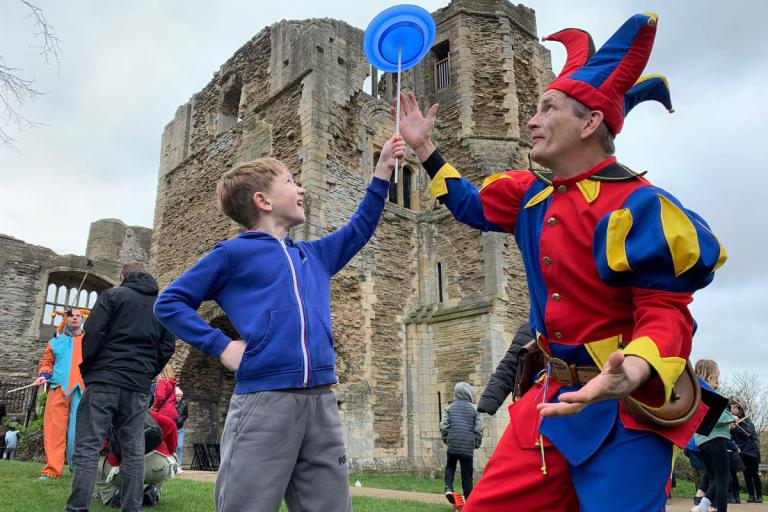 A jester dressed in a bright red, blue, and yellow costume performs a plate-spinning trick with a child in a blue hoodie at Newark Castle. 