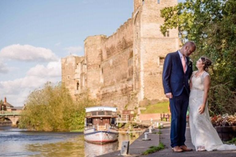 A couple in wedding attire stands on a riverside path near Newark Castle. 
