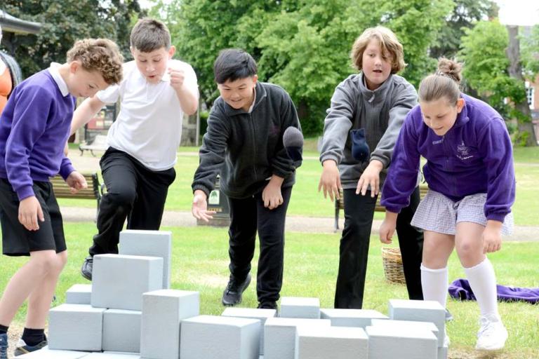 Five excited school children playing an interactive game with large building blocks outside.