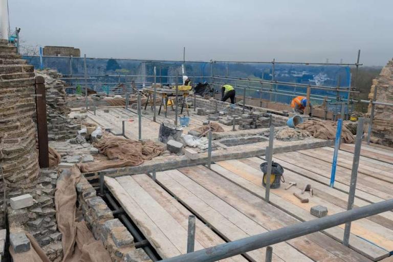 Masons laying straight, level stone courses over uneven medieval masonry on the Gatehouse roof, with scaffolding and materials around the work area.