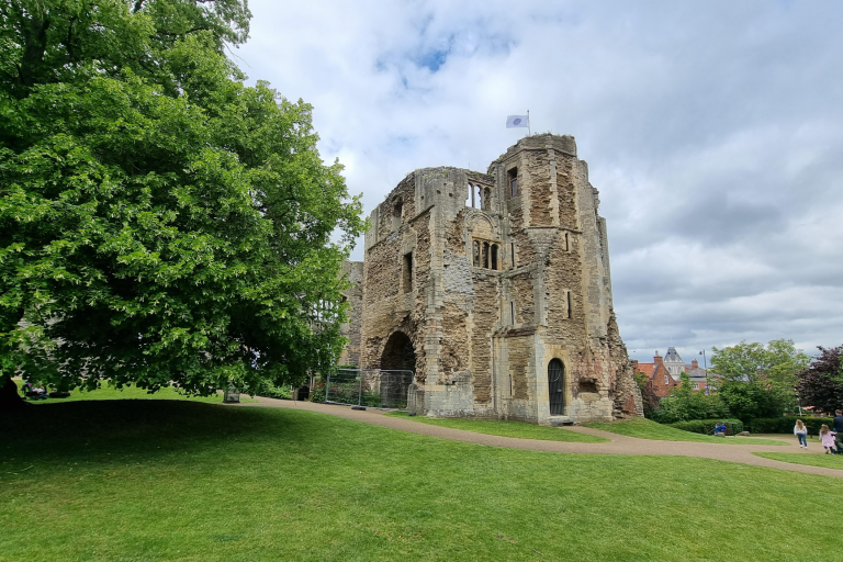 Historic stone castle gatehouse set within landscaped grounds, with a large tree, walking path and cloudy sky.