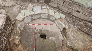 Large worn millstone covering a medieval cesspit, surrounded by stonework, with measuring rods placed across the surface.