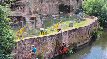 Masons suspended over the river repair Newark Castle’s riverside wall using ropes and safety gear.