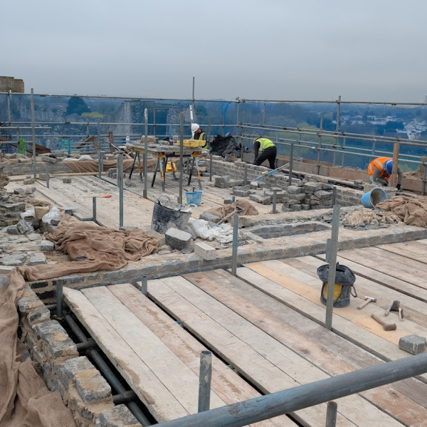 Masons on a high scaffolding platform repair and rebuilding. The area is covered with wooden planks, tools, and building materials, with partially restored stone walls surrounding the site. A hazy landscape with trees and buildings is visible in the background under an overcast sky.